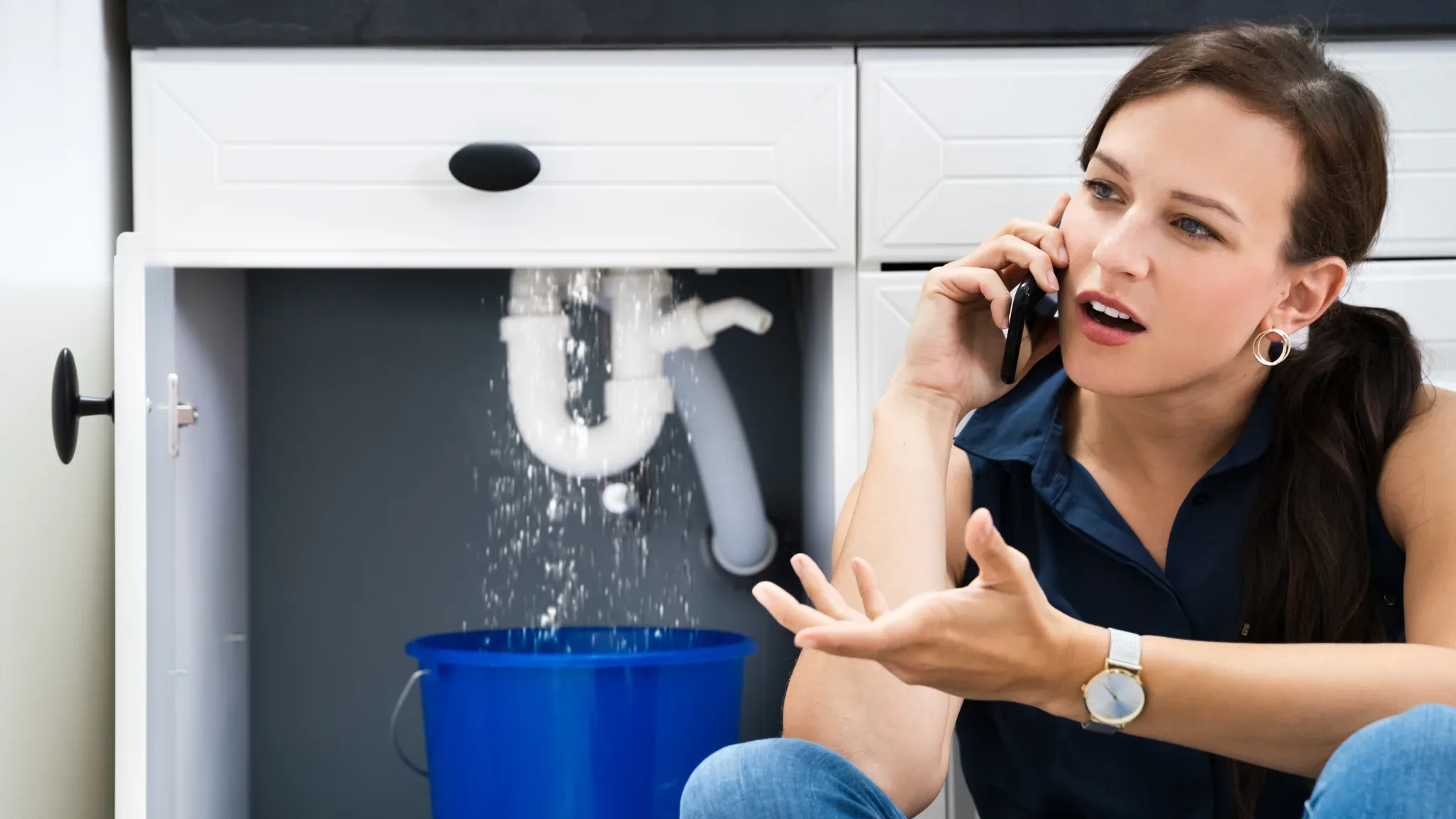 Frustrated homeowner calls a plumber while water leaks from a cabinet, illustrating the urgent need for professional plumbing