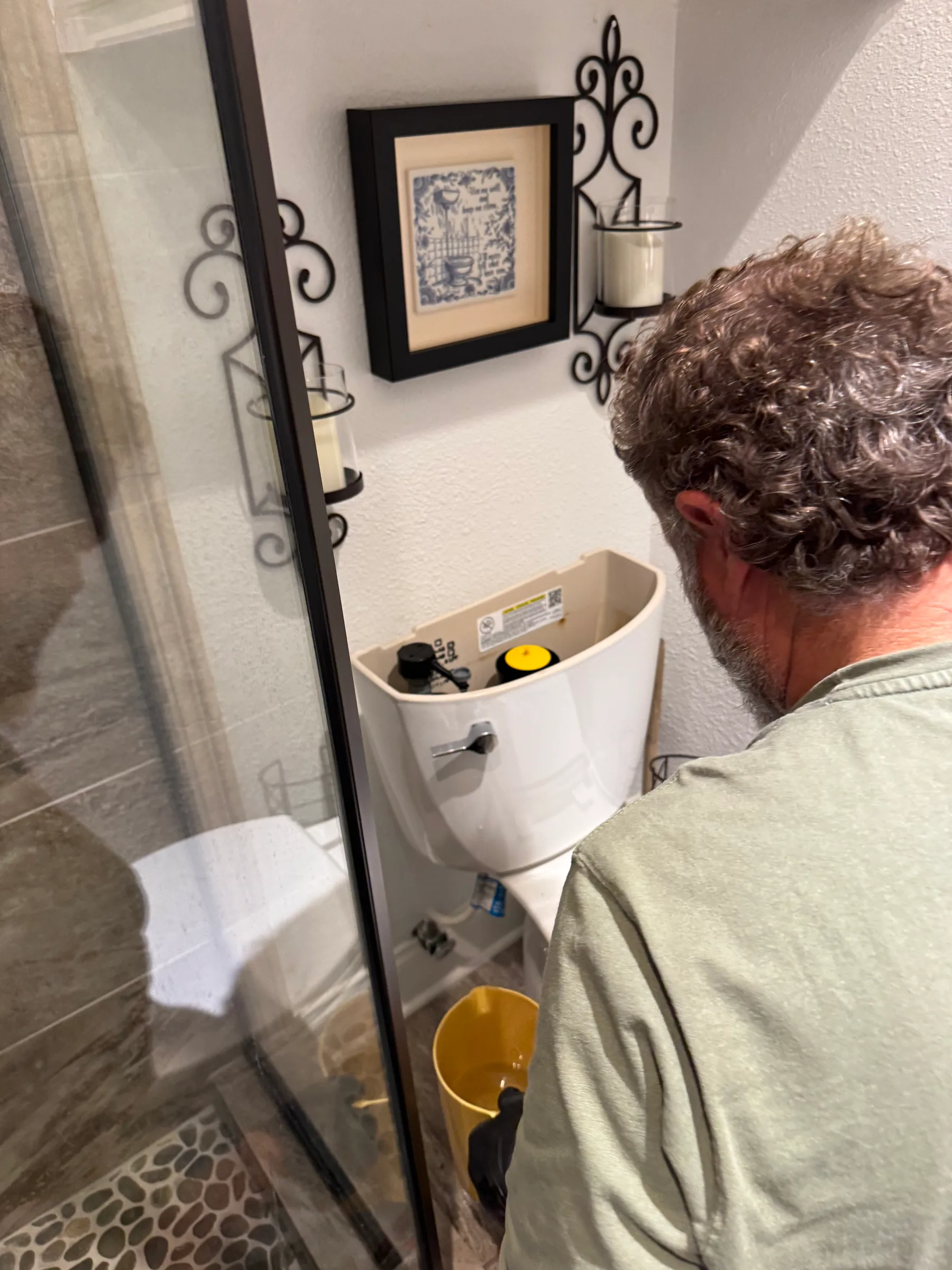 Man inspecting a white toilet in a bathroom with decorative wall sconces and framed artwork.