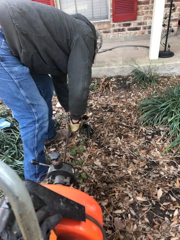 Person using a motorized auger to clear a clogged drain in a residential flower bed.