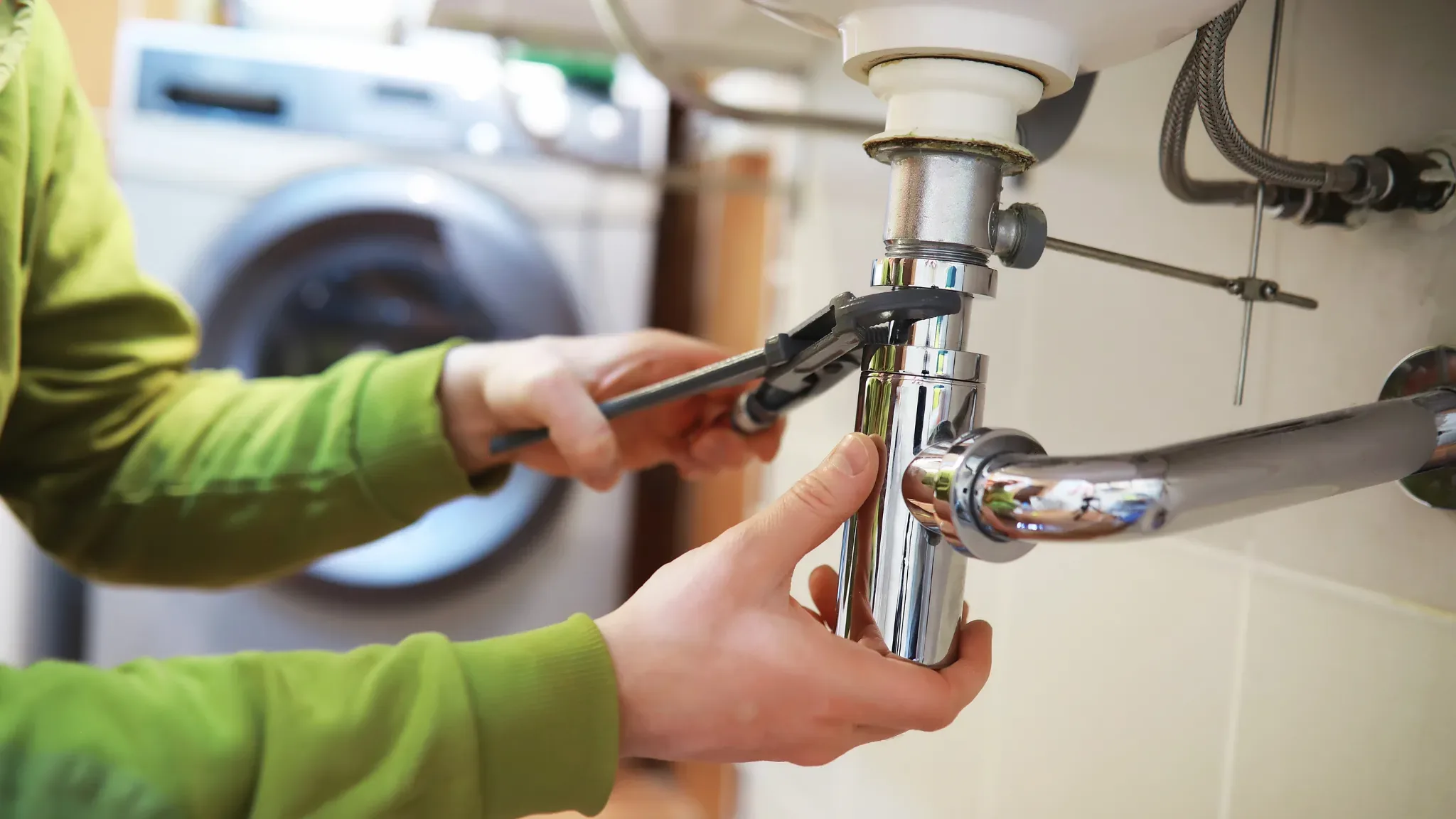 A plumber in a green shirt uses a wrench to repair an under-sink P-trap fixture in a residential bathroom.