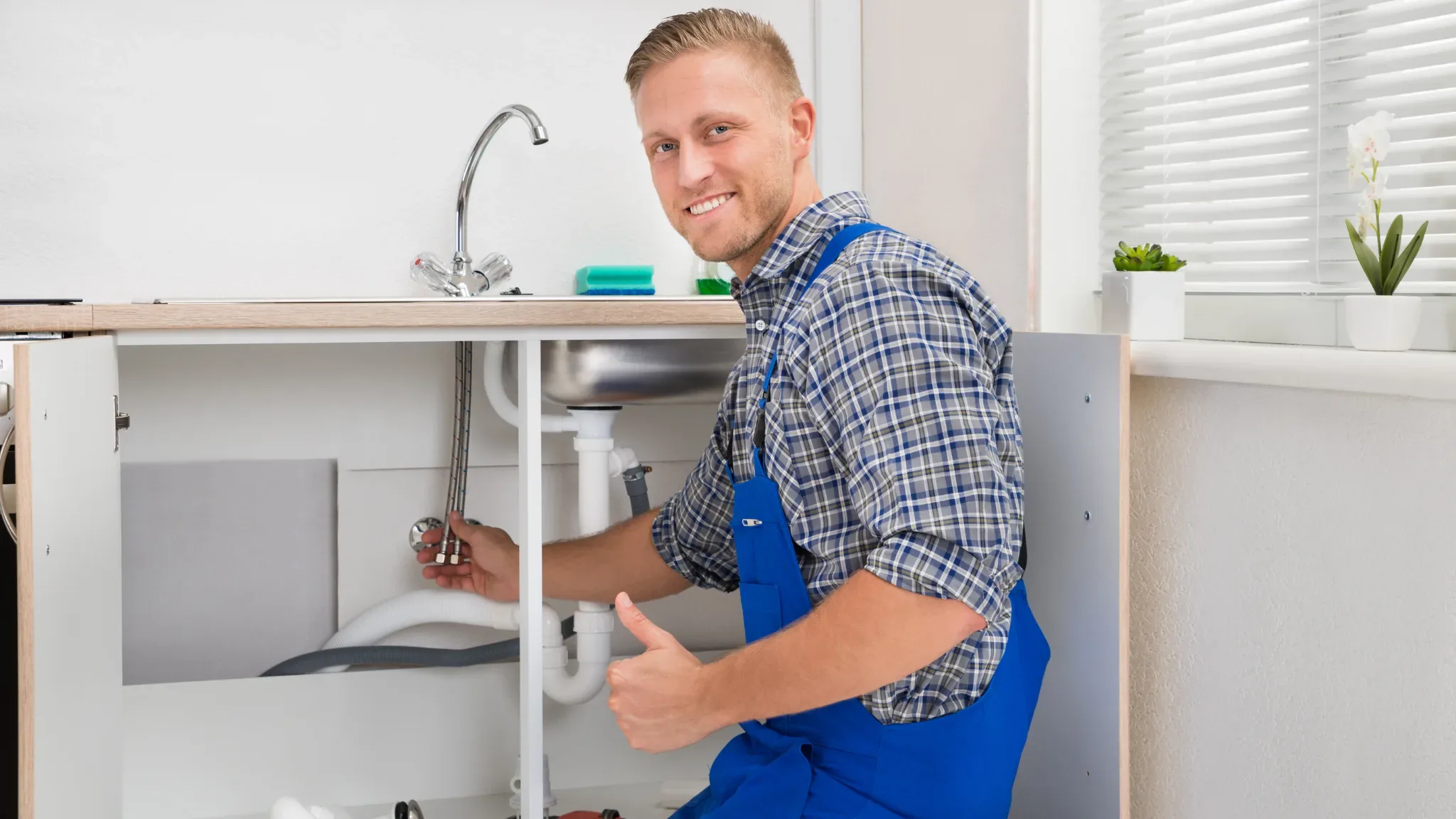 A smiling plumber in blue overalls gives a thumbs up while working under a kitchen sink, conveying expertise and customer sat